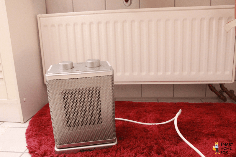 Der Heizlüfter steht auf einem Teppichvorleger neben einer Heizung im Bad und das Stromkabel liegt daneben.