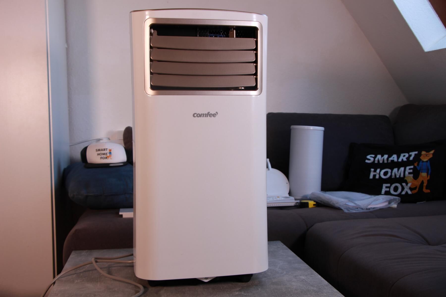 Die Comfee mobile 7000 Easy Cool 2.0 mobile Klimaanlage steht auf einem Couchtisch vor einer Couch und einem Schrägdach Fenster..