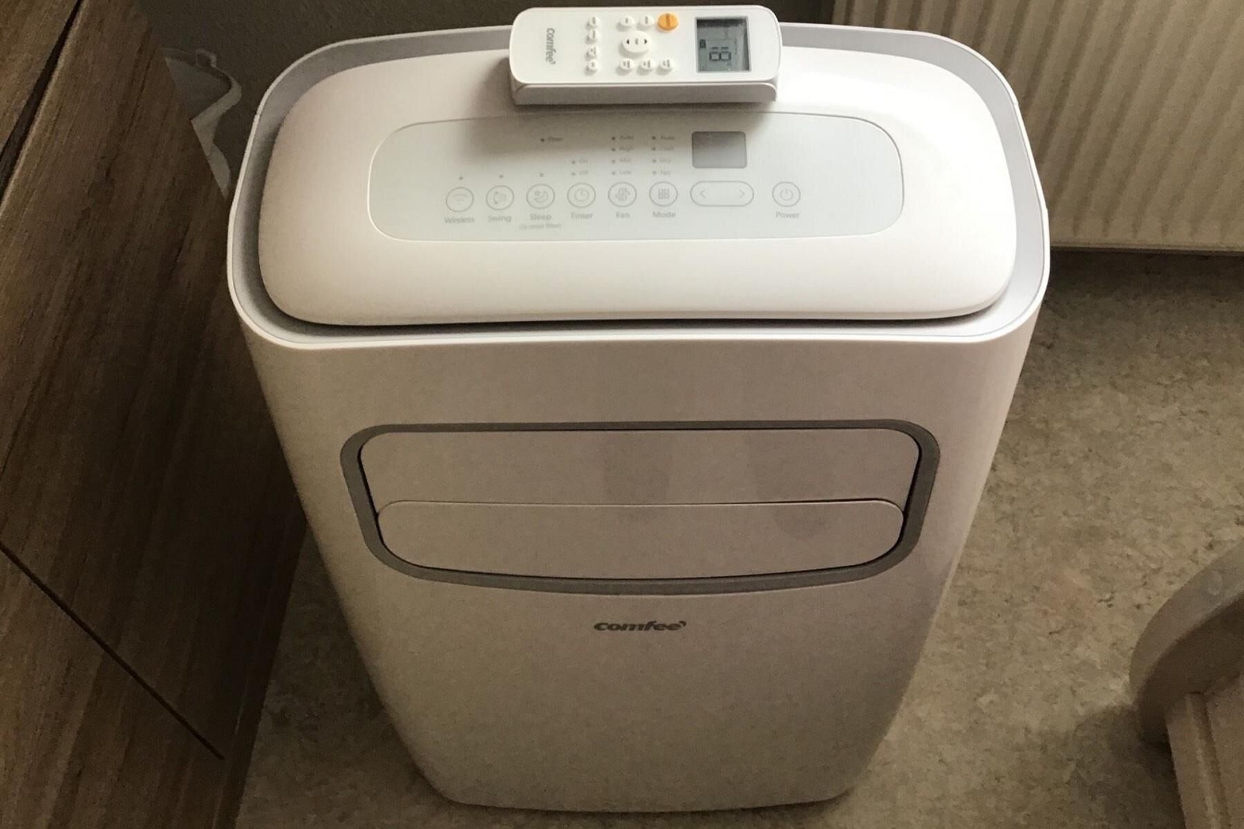 Die Comfee PAC 12000 mobile Klimaanlage steht auf dem Boden vor einer Heizung und die Fernbedienung liegt drauf.