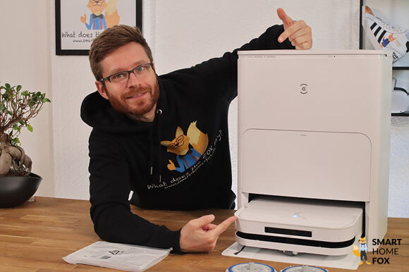 ECOVACS DEEBOT X5 OMNI steht samt Station auf dem Tisch und ein Smart Home Mitarbeiter zeigt mit den Fingern auf den Roboter.