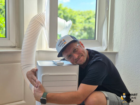 Smart-Home-Fox Experte umarmt mobile Monoblock-Klimaanlage mit Abluftschlauch im Schlafzimmer beim Testen.