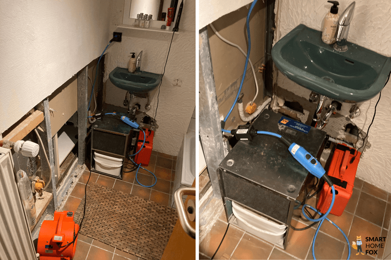 In einem Badezimmer ist die Wand aufgerissen und ein Bautrockner steht vor der offenen Wand unter einem Waschbecken und ein Bauventilator ist daran angeschlossen, um trockene Luft in Richtung des feuchten Mauerwerks zu blasen.