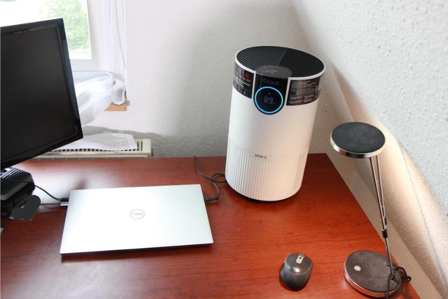 Der Shark HP100EU HEPA Luftreiniger steht eingeschaltet auf einem Schreibtisch neben einem Laptop und das Display des Luftreinigers leuchtet blau.