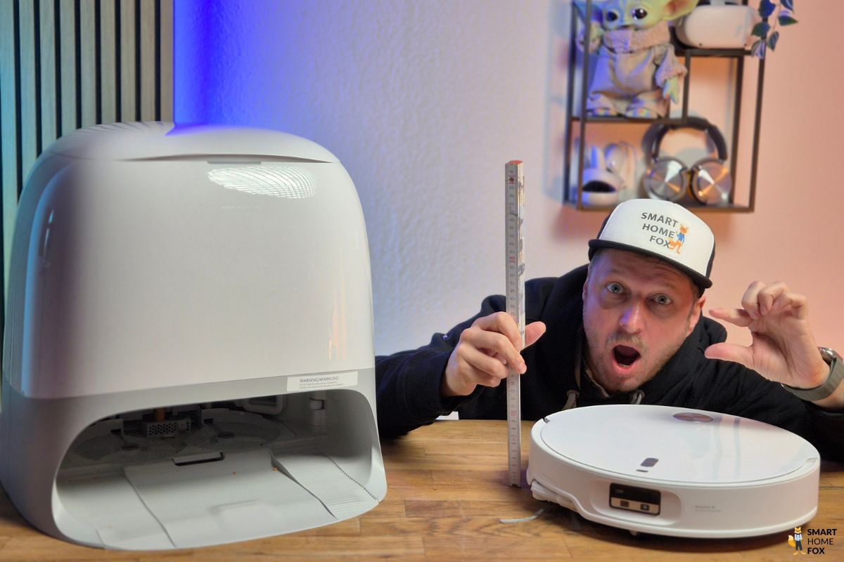Der Roborock Qrevo Curv 2 Pro steht samt Absaugstation im Test bei Smart-Home-Fox auf dem Tisch und wird vom Experten mit einem Zollstock in der Höhe gemessen und der Experte ist sehr überrascht.