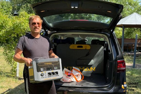 Die VTOMAN FlashSpeed 1500 Powerstation im Test, wird vom Smart-Home-Fox Experten aus dem Auto gehoben, die Anschlüsse auf der Front sind zu sehen und das Solarpanel ist im Hintergrund im Autokofferraum zu sehen.